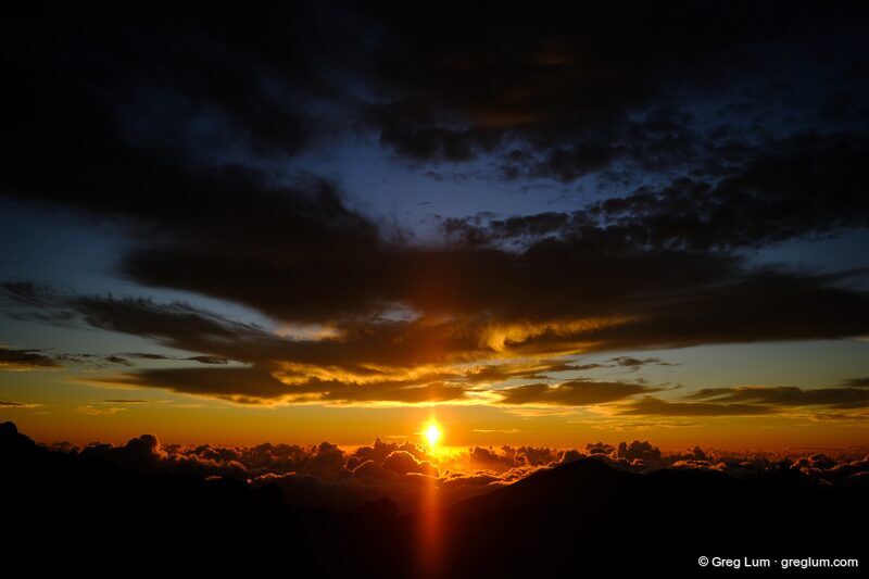 The sun rising over a sea of clouds, under dark streaks of cloud, with a ridge in silhouette.
